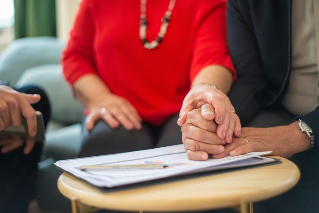 Ein älteres Ehepaar hält Händchen über einem Tisch mit Dokumenten – ein Symbol der Unterstützung im Büroalltag.