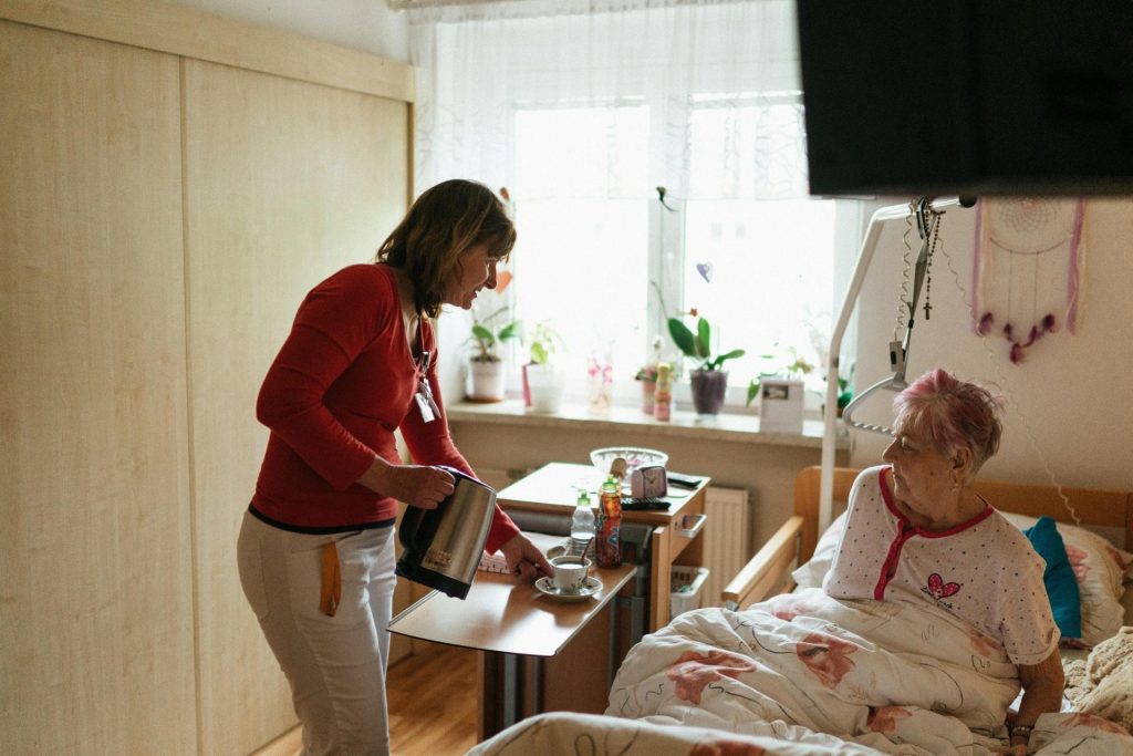 Eine Pflegekraft serviert einer älteren Dame in einem gemütlichen Schlafzimmer Tee und vermittelt dabei Geborgenheit und Fürsorge.