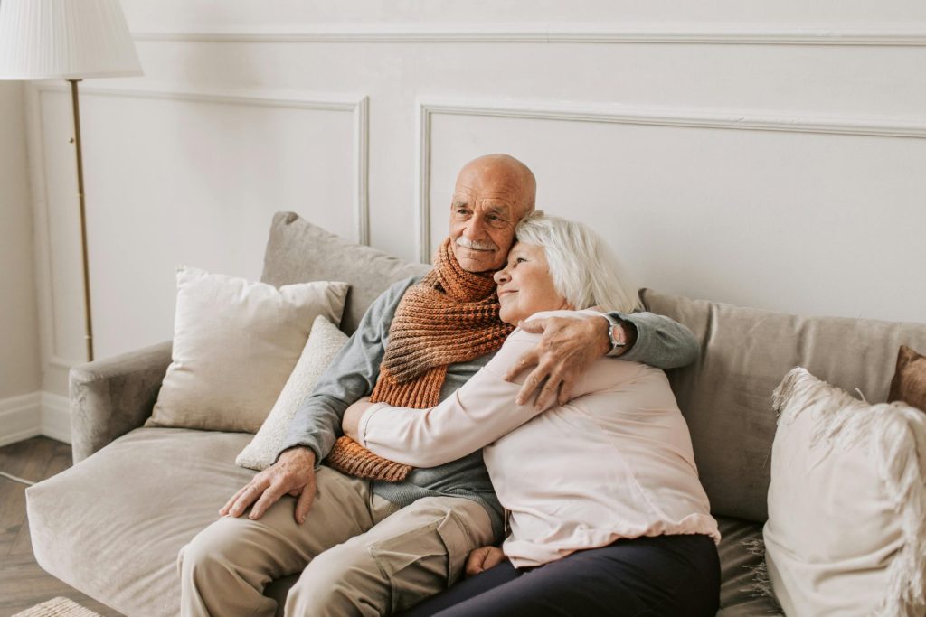 Ein älteres Paar umarmt sich liebevoll auf einem Sofa und symbolisiert damit beständige Liebe und Verbundenheit.